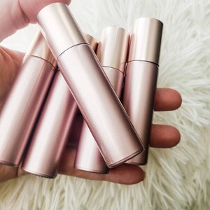 May include: Five rose gold roller bottles with silver caps. The bottles are grouped together and held in a hand.