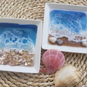 May include: Two white square ceramic dishes with resin ocean scenes. The dishes feature blue water, white foamy waves, and brown sand with seashells. One dish has a larger seashell in the center.