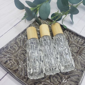 May include: Three clear glass roller bottles with bamboo lids on a gray patterned surface. The bottles are empty and ready to be filled with essential oils or other liquids.