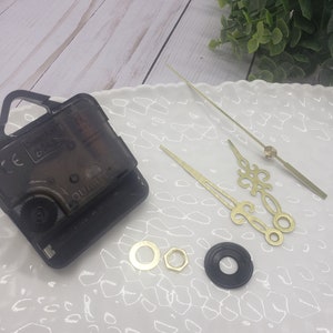 May include: Clock movement kit with black clock mechanism, gold clock hands, and two gold mounting hardware pieces on a white surface.