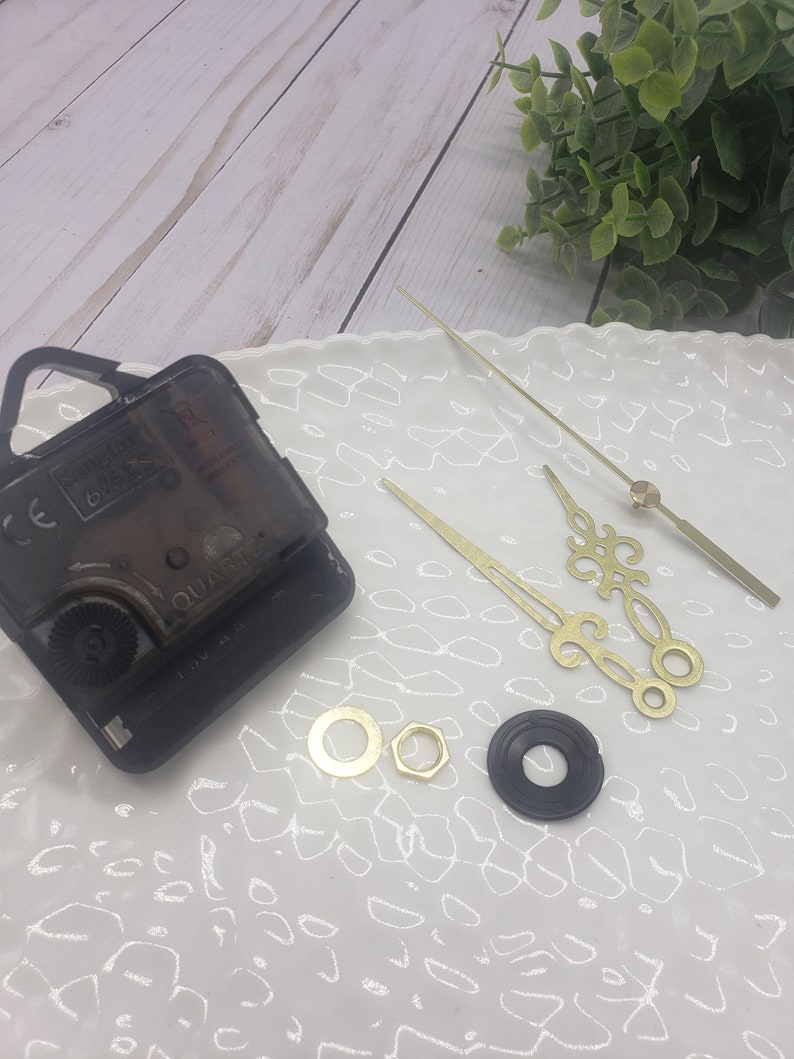 May include: Clock movement kit with black clock mechanism, gold clock hands, and two gold mounting hardware pieces on a white surface.