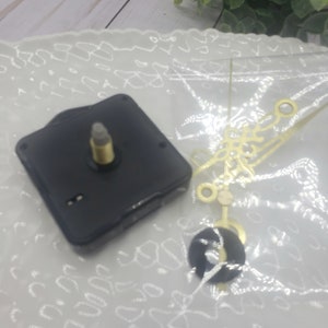 May include: A black clock movement with a gold-colored clock hand set. The clock movement is on a white surface with a lace pattern.