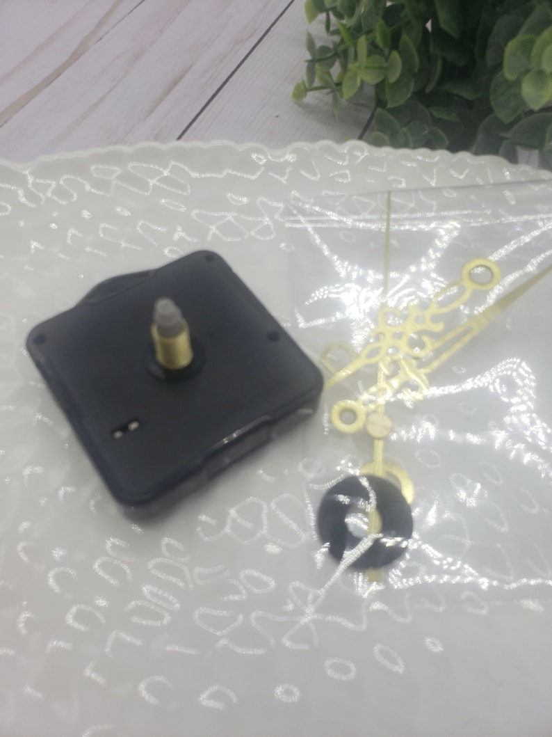 May include: A black clock movement with a gold-colored clock hand set. The clock movement is on a white surface with a lace pattern.