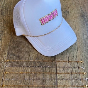 May include: Five gold and silver chain straps with clasps. The straps are laid out on a wooden surface. A white trucker hat with a pink "Howdy" patch is in the background.