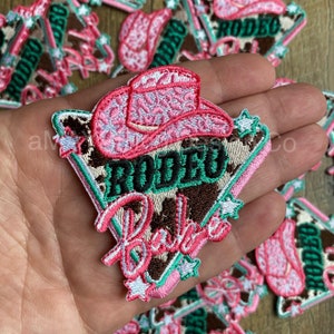 May include: Pink and green embroidered iron-on patch with a cowboy hat, stars, and the text "Rodeo Babe".