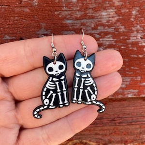 May include: A pair of black cat skeleton earrings with silver hooks. The cat skeletons are white on a black background.