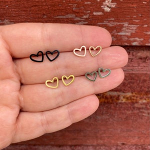 May include: Four pairs of heart-shaped earrings in different colors: black, gold, silver, and rose gold. The earrings are all the same size and shape, and they are shown on a hand.