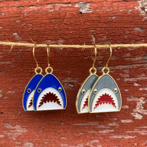 May include: A pair of gold shark earrings. One pair is blue and white, the other is gray and white.