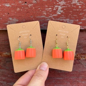 May include: Two pairs of orange and green pumpkin-shaped earrings on brown cards. The earrings have silver and gold hooks. The cards have the text "aMAYSEing Design Co."