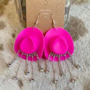 May include: Pink cowboy hat shaped earrings with silver chains and rhinestones. The earrings are hanging from a brown card with the text "aMAYSEing Design Co."