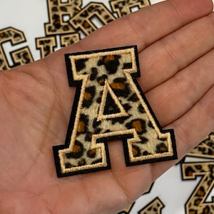 May include: A large, felt letter A in a leopard print pattern with a gold outline. The letter is on a hand.