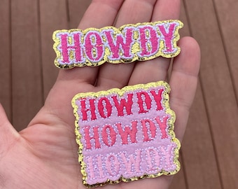 May include: Two pink and gold glitter iron-on patches with the word "Howdy" embroidered in pink. The top patch has the word "Howdy" written once, and the bottom patch has the word "Howdy" written three times, stacked on top of each other.