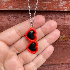 May include: A silver chain necklace with a red heart-shaped sunglasses pendant. The sunglasses have black lenses.