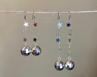 May include: Silver earrings with a disco ball charm and a dangling chain of stars and crescent moons. The earrings are on a string.