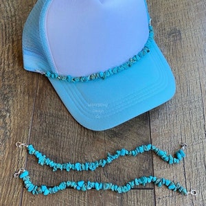 May include: A light blue trucker hat with a white mesh back and a turquoise stone trim. Two matching turquoise stone chains are below the hat. The hat has the text "bMAYSEing Design Co."