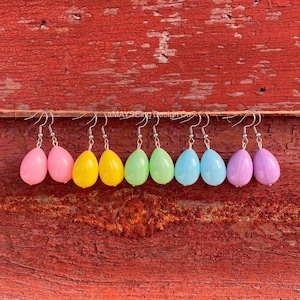 May include: A set of pastel colored egg-shaped earrings. The earrings are made of plastic and have silver hooks. The colors of the earrings are pink, yellow, green, blue, and purple.