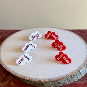 May include: A collection of heart-shaped acrylic charms in red and white, arranged on a wooden slice. The charms feature phrases like "LOVE," "XOXO," and "BE MINE" in red lettering, perfect for Valentine's Day or romantic gifts.