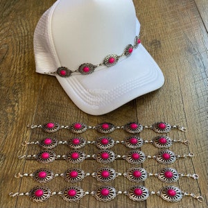 May include: Set of six silver-tone chain bracelets with pink oval-shaped stones. Each bracelet has a floral design around the stones. The bracelets are shown on a wooden surface with a white trucker hat with a silver-tone chain with pink stones on top.