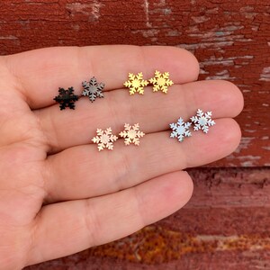 May include: Six snowflake earrings in different colors: black, gold, silver, and rose gold. The earrings are displayed on a hand.