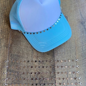 May include: A light blue trucker hat with a white mesh back and a silver star chain trim. The hat is laying on a wood surface with several silver star chains laying next to it.