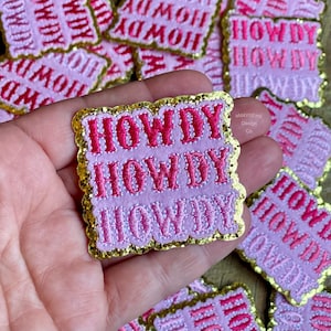 May include: Three pink and gold embroidered patches with the word "HOWDY" in a stacked design. The patches have a glittery finish and a scalloped edge.