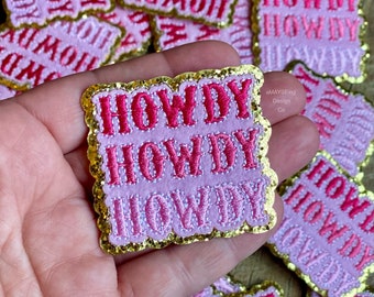 May include: Three pink and gold embroidered patches with the word "HOWDY" in a stacked design. The patches have a glittery finish and a scalloped edge.