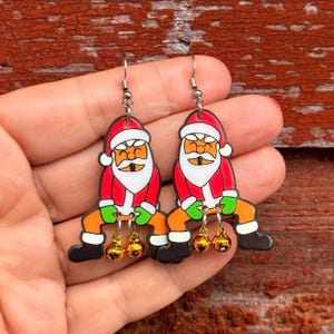 May include: A pair of dangle earrings featuring a cartoon Santa Claus character wearing a red hat, red coat, and green gloves. Each earring has two small gold-colored jingle bells attached to the bottom. The text aMAYSEing Design Co is visible.