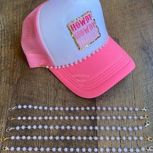 May include: A pink and white trucker hat with a gold glitter patch that says "Howdy Howdy Howdy". The hat is decorated with a row of white pearls around the brim. Five strands of white pearl necklaces with gold clasps are shown below the hat.