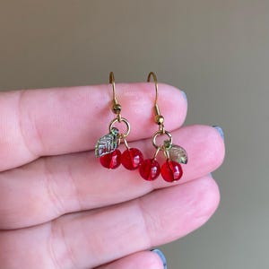 May include: Gold dangle earrings with red glass cherry charms and green leaf accents.