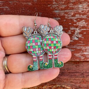 May include: A pair of silver dangle earrings featuring a green, red, and white glitter disco ball with reindeer antlers and elf legs. The earrings are on a hand.