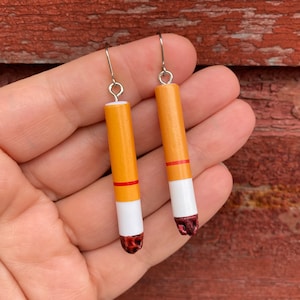 May include: A pair of dangle earrings shaped like cigarettes. The earrings are made of yellow and white clay with a red stripe. The earrings are hanging from silver hooks.