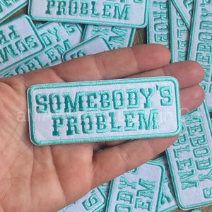 May include: White embroidered patch with a teal blue border. The patch says "Somebody's Problem" in a bold, blocky font.