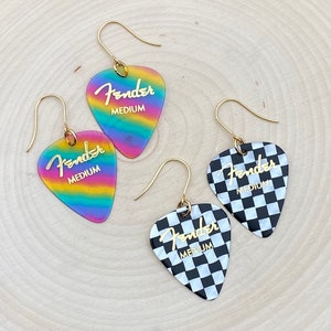 May include: A pair of gold-tone earrings featuring Fender guitar picks. One pick is rainbow iridescent and the other is black and white checkered.