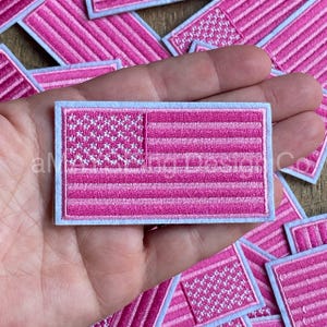 May include: A pink and white embroidered American flag patch with a white border. The patch is held in a hand.