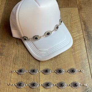 May include: Three silver chains with black oval-shaped stones set in a floral design. The chains are on a wooden surface. A white trucker hat with a mesh back is in the background with the same chain wrapped around the hat.