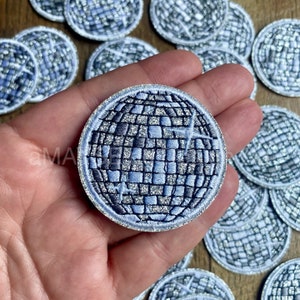 May include: A silver and blue glitter disco ball iron-on patch. The patch is round and has a silver glitter outline. The disco ball is made of many small squares.