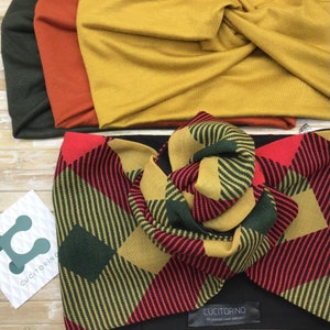 Op de afbeelding: Een rode, groene en gouden gestreepte turban met een knoop bovenop. De turban is gemaakt van een zachte, rekbare stof. De merknaam "Cucitorino" staat gedrukt op een zwart label dat aan de turban is bevestigd.