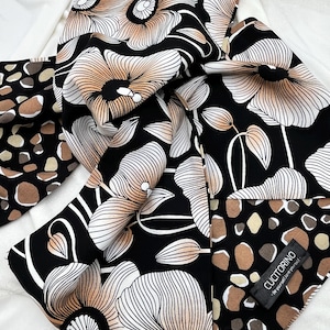 May include: A black and white floral scarf with a repeating pattern of large flowers in shades of peach and white. The reverse side features a pattern of brown and tan organic shapes. The scarf is folded on a white surface.