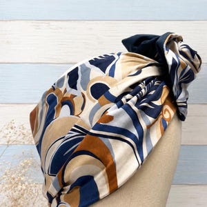 Pode incluir: Um turbante com um padrão abstrato bege, azul marinho e castanho. O tecido é enrolado e amarrado no topo, com um detalhe azul marinho liso. O adorno de cabeça é exibido em uma cabeça de manequim.