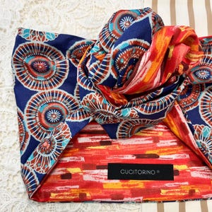 Puede incluir: Una diadema de tela con base azul oscuro y un patrón repetitivo de motivos circulares tipo mandala en naranja, rojo y azul claro. La diadema incluye una sección superior anudada y una etiqueta negra rectangular con el texto CUCITORINO.