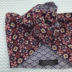 May include: A rolled-up necktie with a repeating pattern of burgundy, navy, and tan floral and geometric shapes. The reverse side features a navy blue and white wave pattern. The label reads "CUCITORINO - be yourself love yourself."