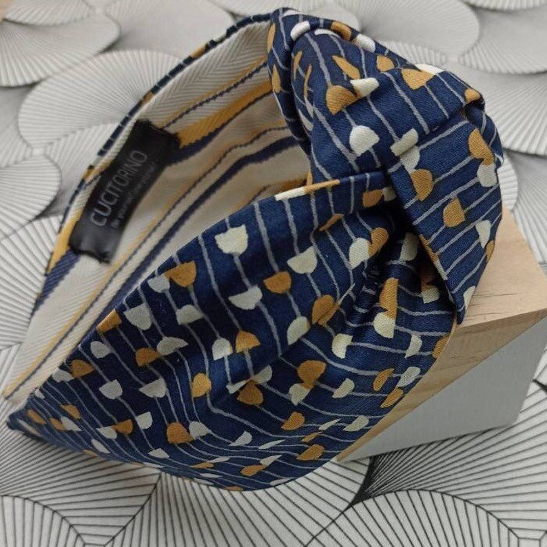 May include: A navy blue knotted headband with a repeating pattern of white and gold half-circles and vertical lines. The interior lining features yellow and white stripes. The label reads "CUCITORINO".