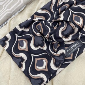 Op de afbeelding: Donkerblauwe haarband met een herhalend geometrisch patroon in wit en beige. De haarband heeft een gedraaide knoop en een klein label met de tekst "CUCITORINO".