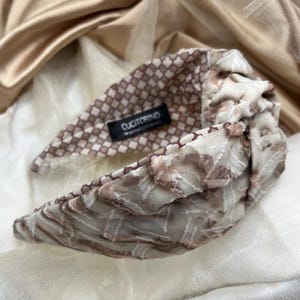 Può includere: Una fascia per capelli in tessuto con un design annodato. La fascia presenta un mix di motivi, tra cui un motivo a scacchi marrone e bianco e un design marmorizzato nei toni del marrone e del bianco. L'etichetta recita "CUCI TORINO".