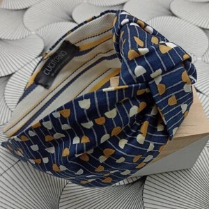 May include: A navy blue headband with a knotted design. The headband features a pattern of white and yellow shapes on a navy background. The interior lining has yellow and white stripes. The label reads "CUCITORINO".