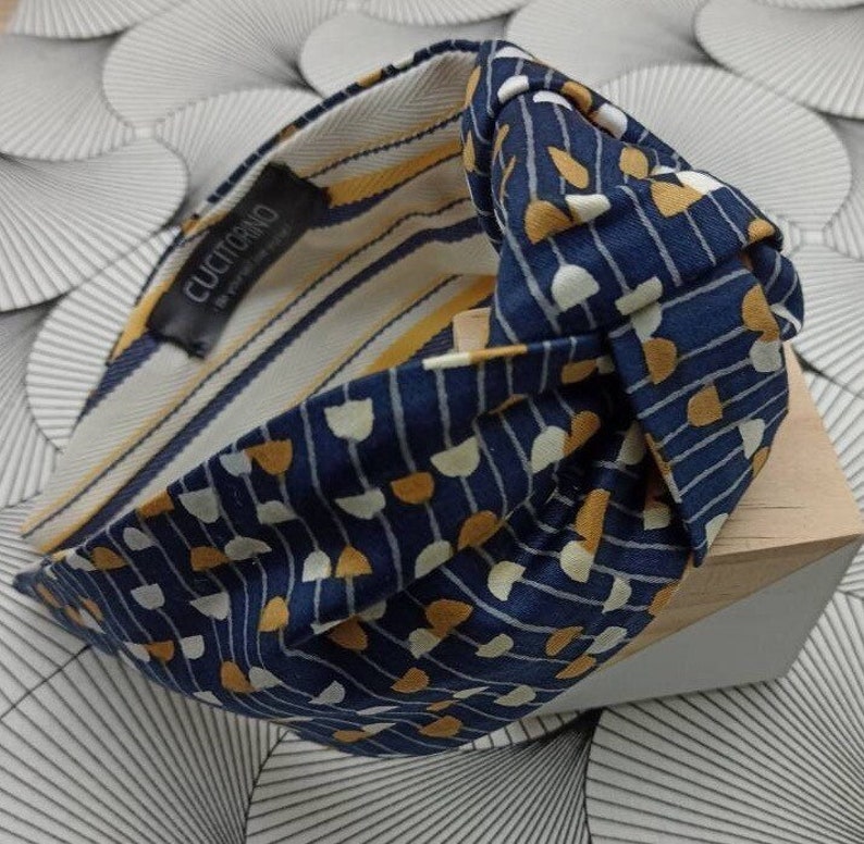 May include: A navy blue headband with a knotted design. The headband features a pattern of white and yellow shapes on a navy background. The interior lining has yellow and white stripes. The label reads "CUCITORINO".