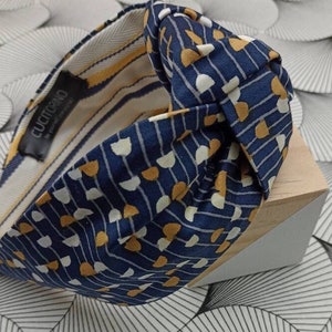 May include: A navy blue headband with a knotted design. The fabric features a pattern of white and gold shapes on a navy background. The interior lining has yellow and white stripes. The label reads "CUCITORINO".