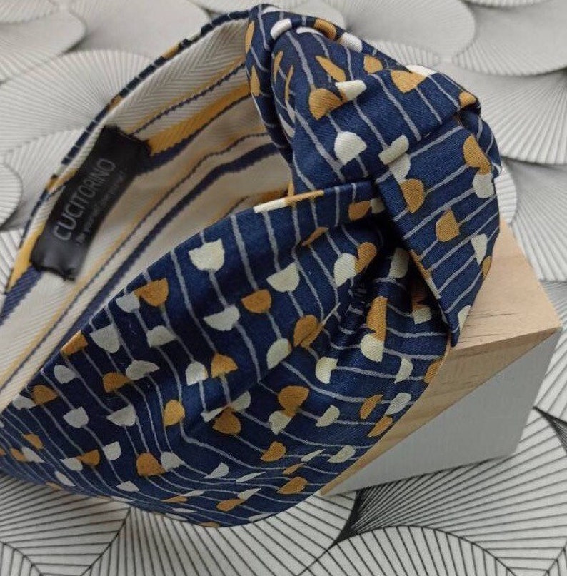 May include: A navy blue headband with a knotted design. The fabric features a pattern of white and gold shapes on a navy background. The interior lining has yellow and white stripes. The label reads "CUCITORINO".