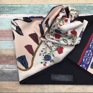 Op de afbeelding: Een beige zijden sjaal met een bloemenpatroon en kwastjes. De sjaal is dubbelgevouwen en heeft een zwarte stoffen achterkant met een label waarop staat "Cucitorino - love yourself, love yourself".
