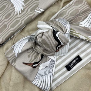 May include: A folded scarf with a crane design in shades of gray, white, and black. The scarf features a striped pattern on one side and a label that reads "CUCITORINO". The scarf is made of a soft, flowing fabric.